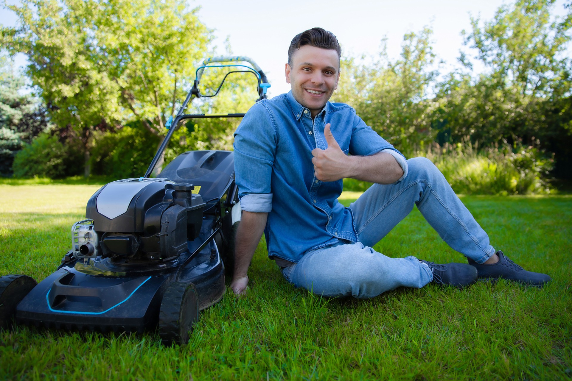 A cheerful man in a denim shirt sits on a lush lawn beside a lawn mower, giving a thumbs-up. The image conveys a sense of accomplishment and satisfaction after completing lawn care on a sunny day.