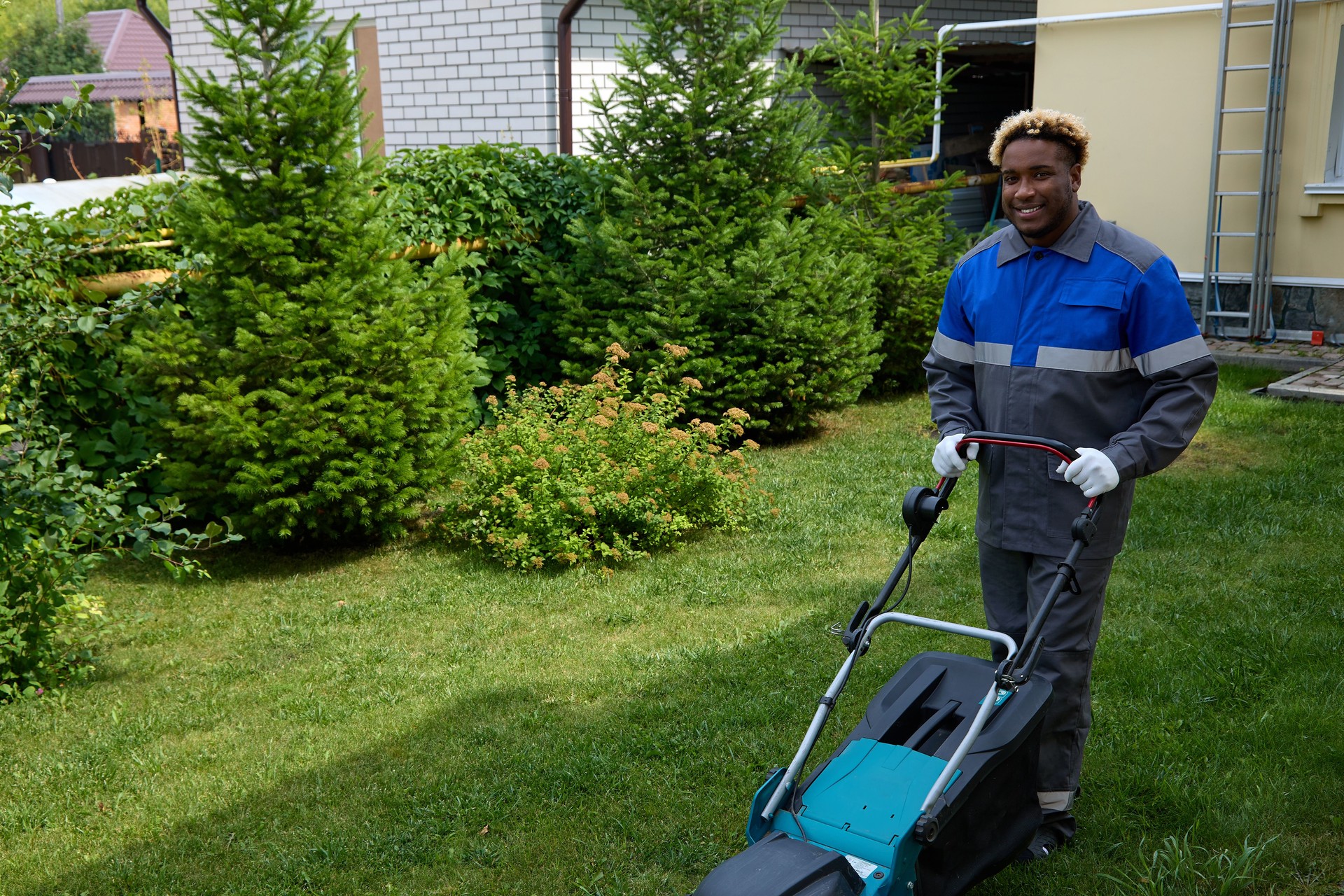 African American man in overalls mows green grass with lawnmower in garden